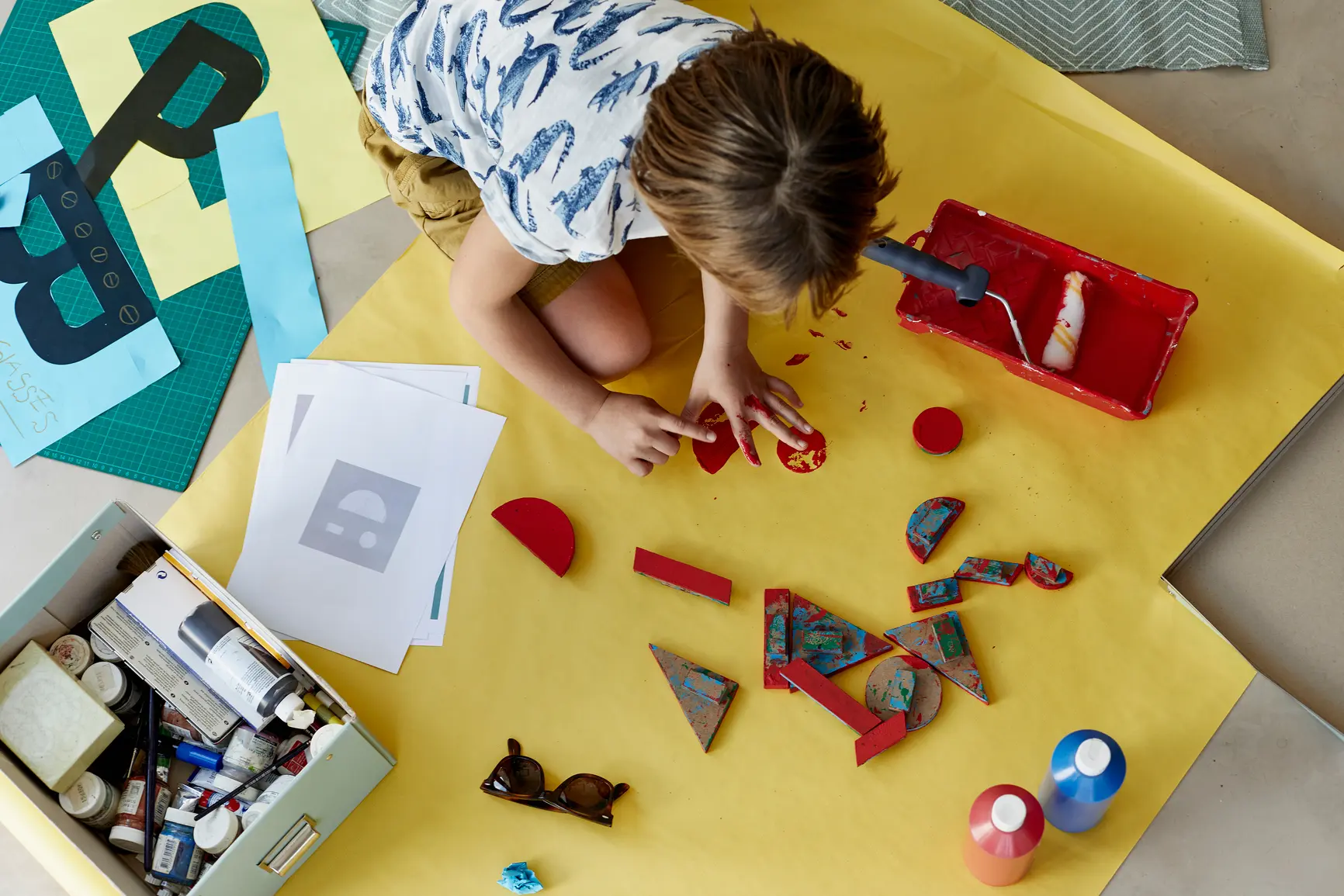 Child engaged in hands-on art activities with geometric shapes and paint on a bright yellow workspace
